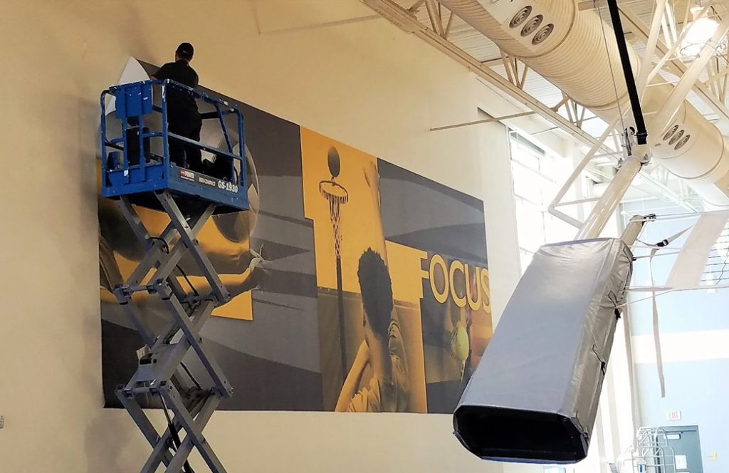 A man on a scissor jack installs a large vinyl wall graphic in a gym.